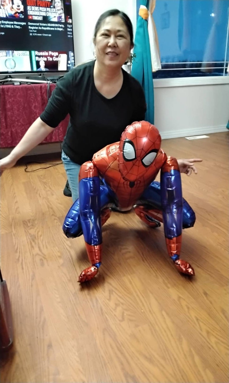 May include: A red, blue, and white Spider-Man balloon in a crouching position. The balloon has a detailed design of the superhero's suit, including the mask and web pattern. The balloon is on a wooden floor.