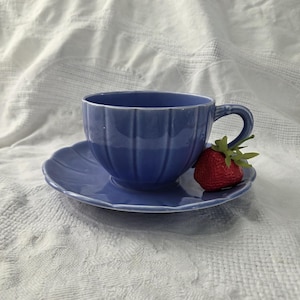 May include: A blue ceramic teacup and saucer set with a ribbed design. A red strawberry with green leaves rests on the saucer. The teacup has a curved handle. The set is on a white textured surface.