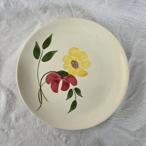 May include: Cream-colored ceramic plate with a hand-painted floral design. The design features a yellow flower, a red flower, and green leaves. The plate is round and has a slightly raised edge.