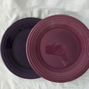 May include: Two round ceramic plates with a concentric ring pattern on the rim. One plate is a deep dark purple, while the other is a lighter shade of plum or magenta. Both plates feature a smooth, glossy finish and a raised circular center detail.