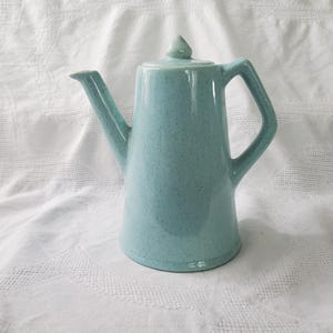 May include: A light blue ceramic teapot with a lid and handle. The teapot has a speckled texture and a spout for pouring. The handle is a square shape. The teapot is on a white surface.