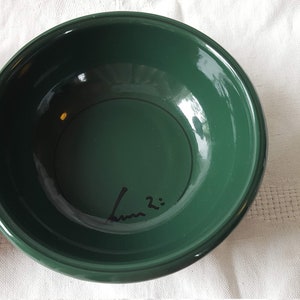 May include: Two ceramic bowls, one red and one dark green, are displayed on a white tablecloth. The green bowl has a handwritten signature and the number 2 on the inside. The bowls are round with a glossy finish.