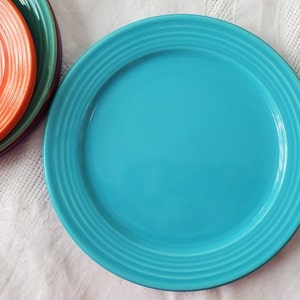 May include: A turquoise blue ceramic dinner plate with a raised rim and concentric circles. The plate is empty and sitting on a white tablecloth.