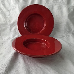 May include: Two circular, glossy red ceramic bowls with wide, flat rims. The bowls feature a deep, recessed centre and a smooth, reflective finish. The design is simple and uniform, showcasing a vibrant, solid red colour throughout the entire surface.