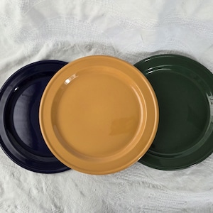Puede incluir: Tres platos de cerámica en fila, cada uno con un color liso diferente. Los platos son azul marino, amarillo mostaza y verde oscuro. Los platos tienen un diseño sencillo y clásico, con un borde ligeramente elevado.