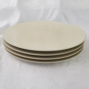 May include: Four beige oval plates stacked together. The plates are a neutral colour and have a simple design. They are placed on a white, textured surface. Each plate's edge is visible.