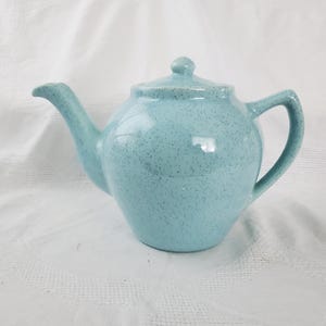 May include: A light blue ceramic teapot with a speckled finish. The teapot has a curved spout, a rounded body, and a handle. The lid has a small knob. The teapot is sitting on a white surface.