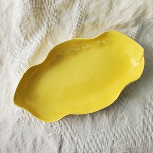 May include: A yellow ceramic serving dish with a wavy edge. The dish is shaped like a leaf and has a smooth, glossy finish.