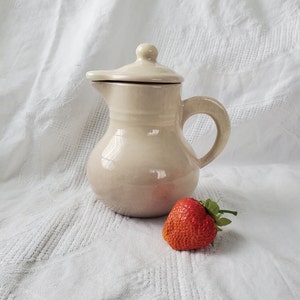 以下が含まれることがあります： 蓋と取っ手がついたクリーム色のセラミックピッチャー。ピッチャーは白い布の表面に置かれ、赤いイチゴの隣にあります。