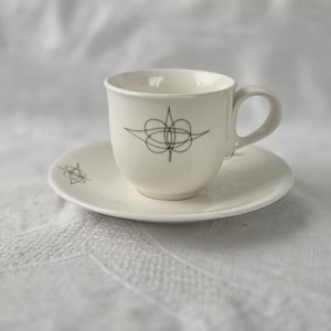 May include: A white ceramic teacup and saucer set. The cup and saucer have a simple black line drawing design. The cup is round with a handle, and the saucer is circular. The set is on a white surface.