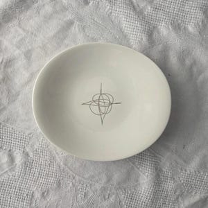 Op de afbeelding: Een witte keramische kom met een zwart abstract ontwerp in het midden. De kom is rond en staat op een wit, getextureerd oppervlak. Het ontwerp is een reeks elkaar kruisende lijnen.