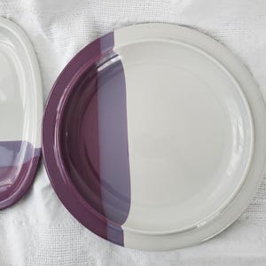 Może przedstawiać: Dwa ceramiczne talerze w paski w kolorze fioletowym i białym. Talerze są okrągłe i mają lekko uniesiony brzeg.