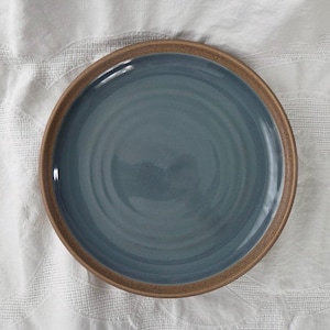 Può includere: Un piatto rotondo in ceramica caratterizzato da un centro liscio e lucido color blu ardesia con sottili solchi circolari concentrici. Il piatto presenta un bordo ampio, non smaltato e testurizzato in una calda tonalità marrone terra.