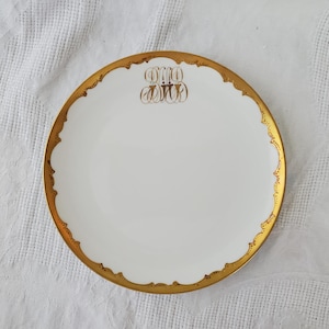 May include: A white ceramic plate with a gold rim and a gold monogram in the center. The monogram is the letters "O.M.O."