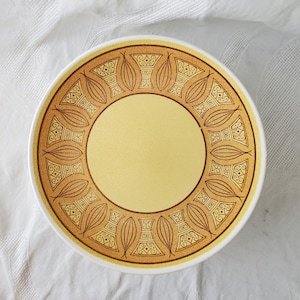 Puede incluir: Un plato grande, redondo, de cerámica blanca con un diseño geométrico marrón y amarillo. El diseño presenta un patrón repetitivo de hojas estilizadas y cuadrados.