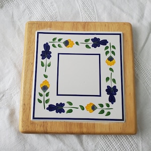 May include: A wooden trivet with a white ceramic tile insert. The tile has a blue and yellow floral design with a blank center.