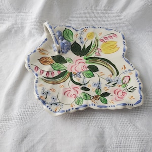 Puede incluir: Un plato de servir de cerámica blanca con forma de hoja y un asa. El plato está decorado con flores pintadas a mano en tonos de rosa, amarillo, verde y azul.