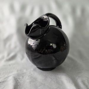 May include: A glossy black ceramic pitcher with a spherical body, a curved handle, and a distinctively ruffled, wavy rim around the pouring spout. The surface is highly reflective, showing light highlights and a smooth, rounded silhouette.