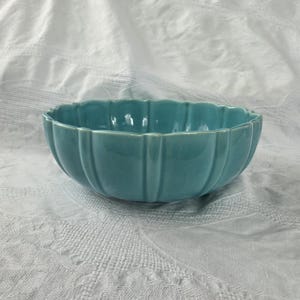 May include: A turquoise ceramic bowl with a scalloped rim. The bowl has vertical ridges and a glossy finish. The bowl is sitting on a white, textured surface.