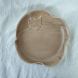 May include: A beige ceramic plate with a leaf design. The plate has a scalloped edge and a raised leaf detail on the rim. The plate is a light brown colour and appears to be handmade.