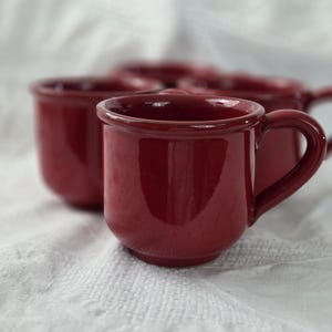 Może przedstawiać: Zestaw czterech błyszczących, głęboko czerwonych ceramicznych kubków. Każdy kubek ma zaokrąglony korpus, mały uchwyt i lekko rozszerzoną krawędź. Kubki są ustawione na białej, teksturowanej powierzchni.