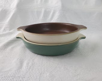 May include: Two stacked ceramic serving dishes. The top dish is brown inside with a cream-colored rim and handles. The bottom dish is a muted green color. Both dishes have a rounded oval shape and are sitting on a white surface.