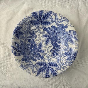 May include: A round, white and blue ceramic plate with a grape and leaf design. The plate features clusters of grapes and detailed leaves in a classic blue and white color scheme. The plate is set against a white background.