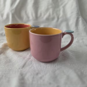 May include: Two ceramic mugs with rounded bodies and handles topped with small, flat, light blue circular accents. One mug is yellow with a pink interior, while the other is pink with a yellow interior, both featuring a smooth, glossy finish.