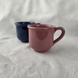 May include: Two small ceramic mugs with a glossy finish. One mug is a dusty rose pink colour and is positioned in the foreground, while the second mug is a deep navy blue colour and is placed slightly behind and to the left of the pink mug.