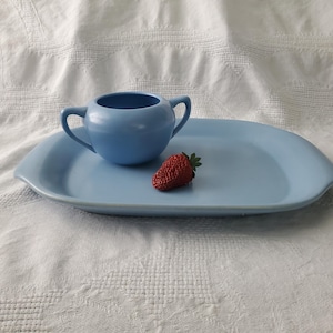 Puede incluir: Un juego de servir de cerámica azul claro con un azucarero pequeño y una bandeja rectangular. Una fresa roja fresca descansa sobre la bandeja. El conjunto se exhibe sobre una superficie blanca texturizada.