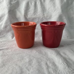 Könnte beinhalten: Zwei Keramikbecher mit einer glatten, glänzenden Oberfläche. Ein Becher ist in einem leuchtenden Orange gehalten, der andere in einem tiefen, satten Rot. Beide Becher haben eine einfache, konische Zylinderform mit einer horizontalen Rille.