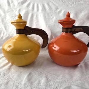 Puede incluir: Dos jarras de cerámica, una amarilla y otra naranja, con asas y tapas de madera. Las jarras tienen forma bulbosa y están sobre un fondo blanco.