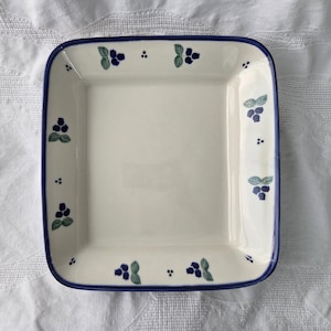 May include: A square ceramic plate with a cream-colored base and a dark blue rim. The border features a repeating pattern of small dark blue berries with light green leaves, interspersed with tiny dark blue dots along the edges.