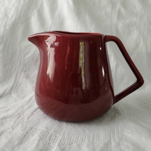 Puede incluir: Una jarra de crema de cerámica de color rojo oscuro con un cuerpo redondeado y un asa que se curva hacia arriba y hacia afuera. La jarra de crema está sobre una superficie blanca.