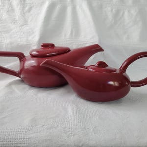 Puede incluir: Dos teteras de cerámica granate con tapas y asas a juego. La tetera más grande tiene un asa en forma de D, mientras que la más pequeña tiene un asa redondeada. Ambas teteras están sobre una superficie blanca texturizada.