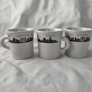 May include: Three white ceramic mugs with black skyline designs. Each mug features a different Seattle landmark, including the Space Needle, Pike Place Market, and the Great Wheel. The mugs have a simple, classic design with a handle.