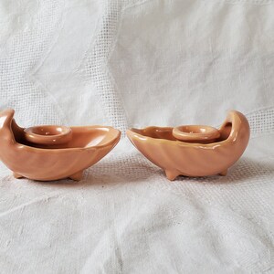 May include: Two peach-colored ceramic candle holders shaped like seashells. Each holder has a small round indentation for a candle.