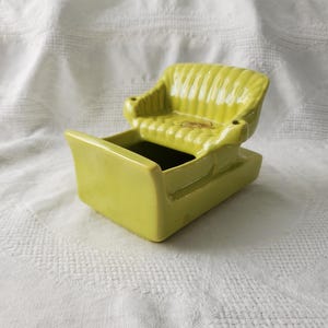 Puede incluir: Una jardinera de cerámica verde lima con forma de silla. La jardinera tiene un asiento pequeño con reposabrazos y una base rectangular. El asiento tiene crestas verticales. La jardinera está sobre una superficie blanca texturizada.