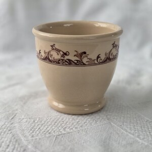 Könnte beinhalten: Eine beige Keramiktasse mit einem braunen floralen Muster am oberen Rand. Die Tasse hat eine abgerundete Form mit einem kleinen Sockel und einem leicht ausgestellten Rand. Der Hintergrund ist eine weiße, strukturierte Oberfläche.