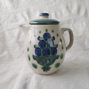 Puede incluir: Una jarra de cerámica blanca con un diseño floral azul y verde. La jarra tiene una tapa y un asa.