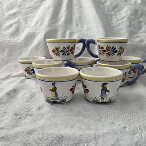 Könnte beinhalten: Eine Sammlung von Keramikbechern und Untertassen mit weißem Grund, gelben Rändern und blauen Griffen. Die Tassen sind mit Blumenmustern in Rot, Blau und Grün verziert, einige zeigen Figuren in traditioneller Kleidung. Das Set ist auf einem weißen Tuch angeordnet.