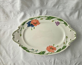 May include: An oval-shaped cream-colored ceramic serving platter featuring decorative handles on each side. The rim is adorned with a painted botanical pattern of orange, blue, and purple poppy flowers connected by green stems and leaves.