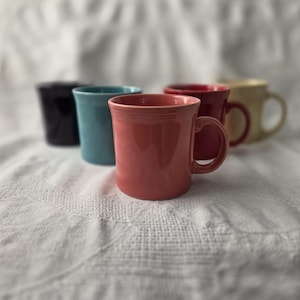 Può includere: Una collezione di cinque tazze in ceramica in vari colori solidi, tra cui corallo, turchese, blu scuro, rosso e crema. Ogni tazza presenta una finitura liscia e lucida, un manico arrotondato e un bordo leggermente svasato.