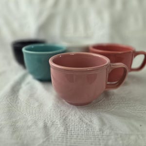 May include: A collection of four ceramic mugs in various colours, including a prominent dusty pink mug in the foreground, followed by a turquoise mug, a dark navy mug, and a white mug, all featuring a smooth finish and a simple handle design.