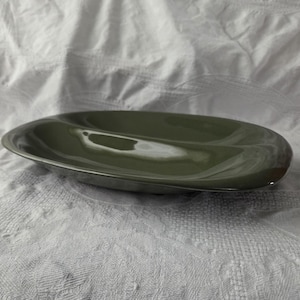 May include: An olive green, oval-shaped serving dish. The dish has a glossy finish and a slightly raised rim. The interior is smooth and reflects light. The dish is designed for serving food.