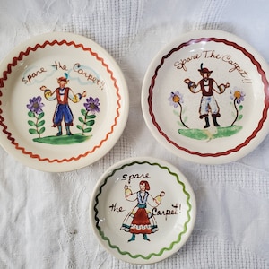 Op de afbeelding: Drie vintage keramische borden met handgeschilderde volkskunstontwerpen. Elk bord toont een centrale figuur in traditionele kledij, omringd door bloemen, met de tekst "Spare the Carpet!!". De borden hebben rood, groene en bruine geschulpte randen.