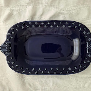 May include: A deep navy blue ceramic serving dish with an oval shape. The wide rim features a repeating pattern of small white stars. On each of the two pointed handles, there is a rectangular design with white horizontal stripes.