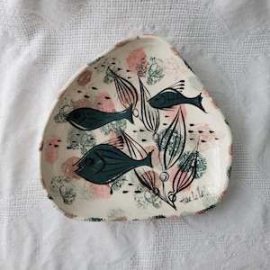 Könnte beinhalten: Eine cremefarbene, dreieckige Keramikschale. Sie zeigt ein handgemaltes Design aus teal-farbenen Fischen, schwarzem Seetang und abstrakten rosa und grünen Formen. Die Schale ist mit "Terre Le Tain" in schwarzer Schrift signiert.