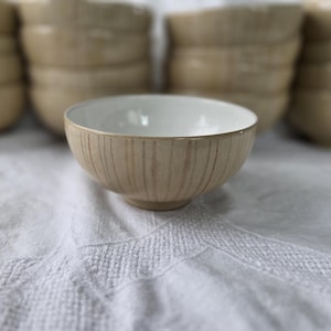 Peut inclure: Un bol en céramique beige avec un intérieur blanc, posé sur un tissu blanc. Le bol présente des lignes verticales texturées à l'extérieur. Plusieurs bols similaires sont empilés en arrière-plan.