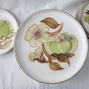May include: A set of white ceramic dinner plates with a floral design featuring green, brown, and pink flowers. The plates have a gold rim.